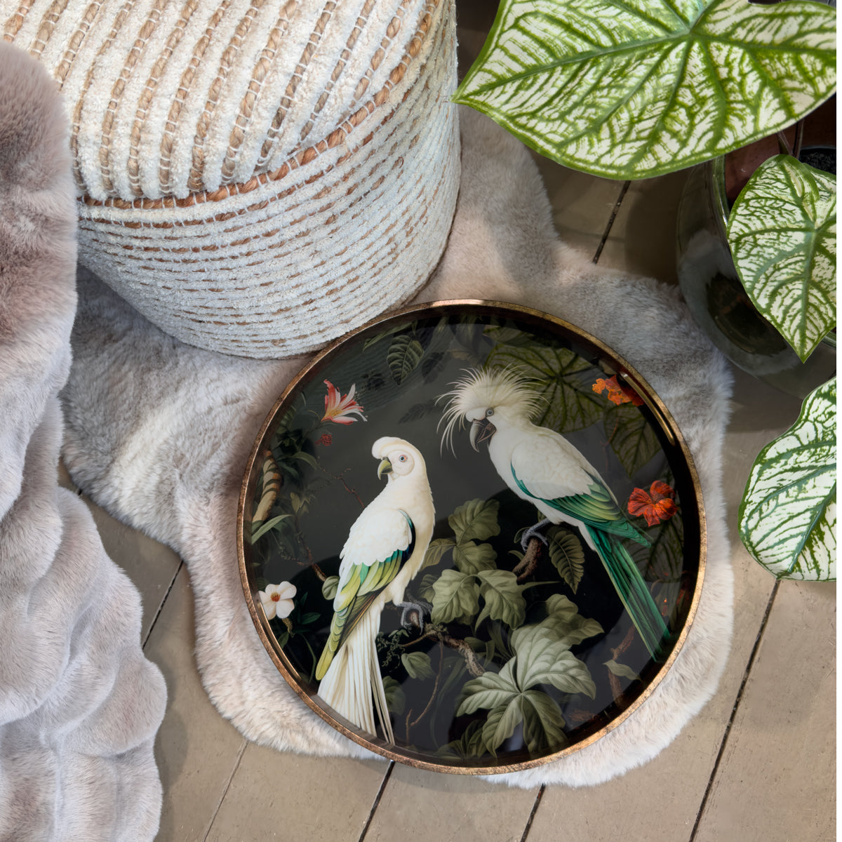 A circular decorative tray featuring two white parrots amidst lush green foliage and red flowers. The tray is placed on a soft faux fur rug beside a textured pouf and large leafy plant.