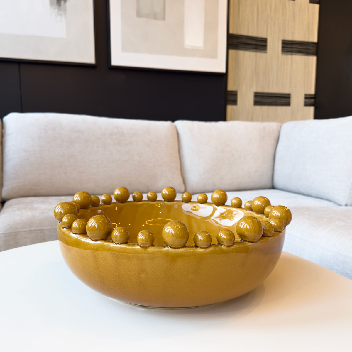 A mustard ceramic decorative bowl sitting on a white coffee table in a living room with pictures on wall.