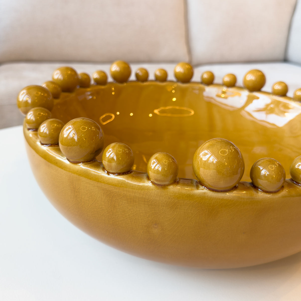 A close up of a mustard coloured bobble surround bowl, sitting infront of a cream sofa.
