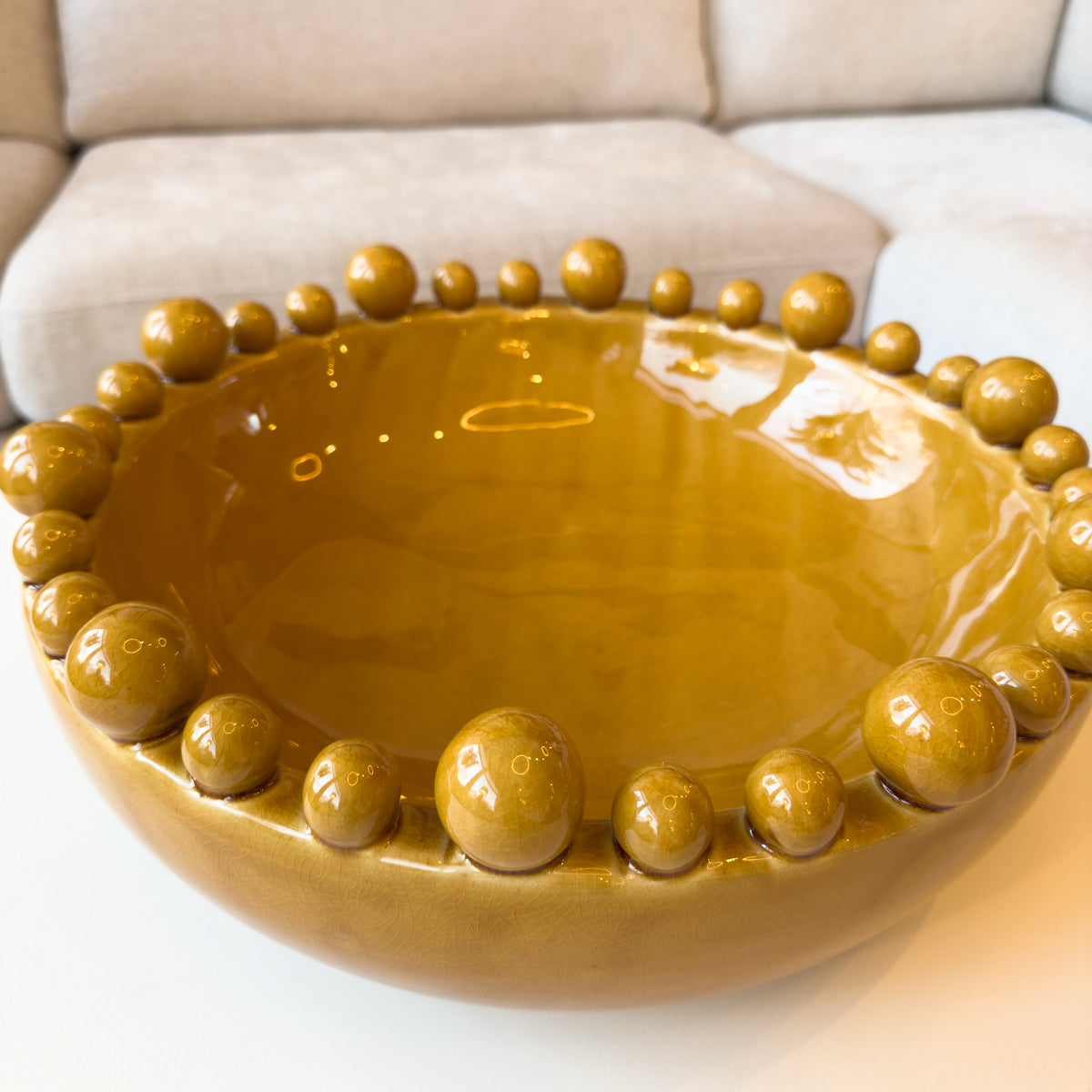 A glossy mustard ceramic bowl with bobble details around the rim, sitting on a white coffee table.