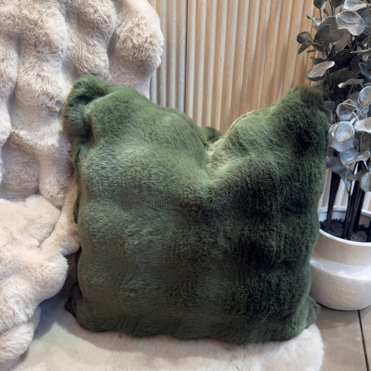 Plush dark green faux fur cushion rests on a cream faux-fur throwbeside a potted plant.