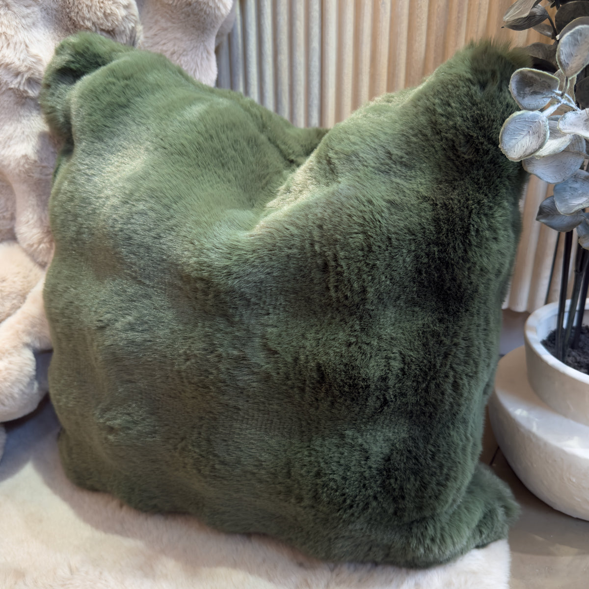 Plush dark green faux fur cushion rests on a cream faux-fur throw beside a potted plant.