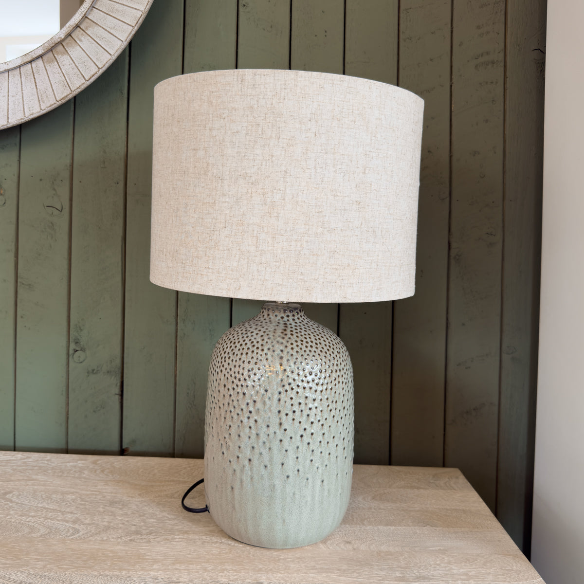 A table lamp turned off, featuring a green base and a cream shade, against a green slatted wooden wall, providing soft illumination for a cozy atmosphere.