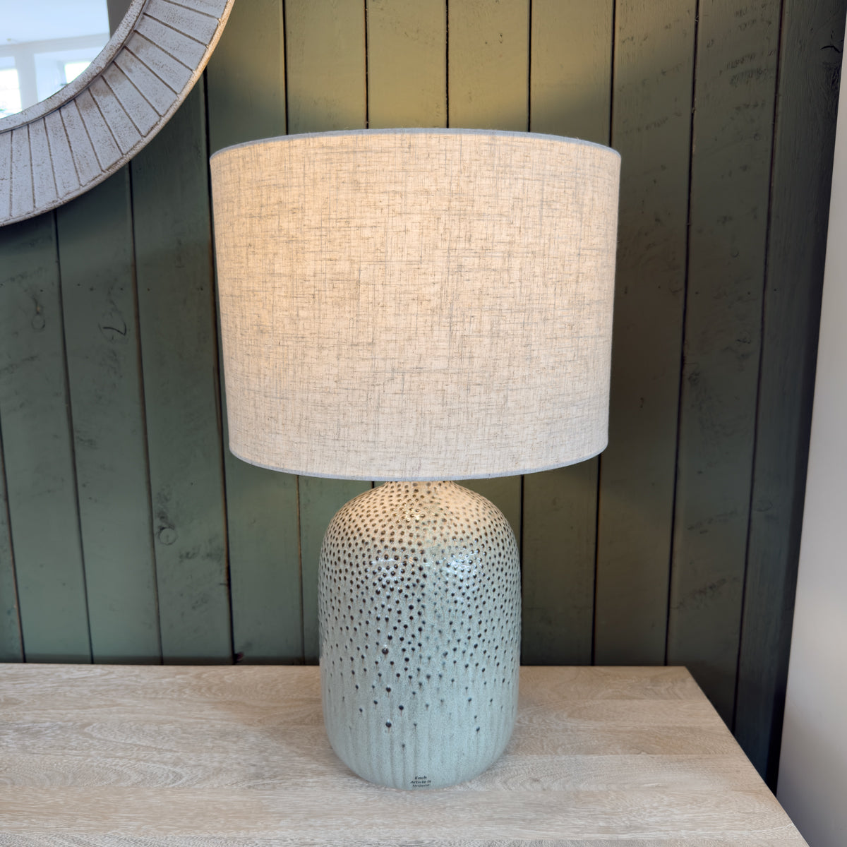 A table lamp featuring a green base and a cream shade, against a green slatted wooden wall, providing soft illumination for a cozy atmosphere.