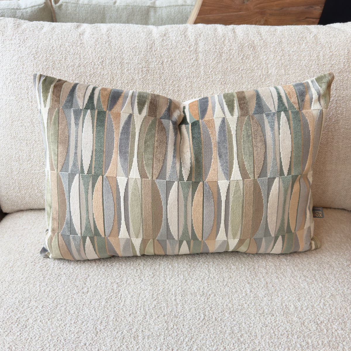 Decorative rectangular pillow with muted geometric leaf patterns in beige, gray, blue, and sage green, resting on a cream sofa; calm, cozy tone.