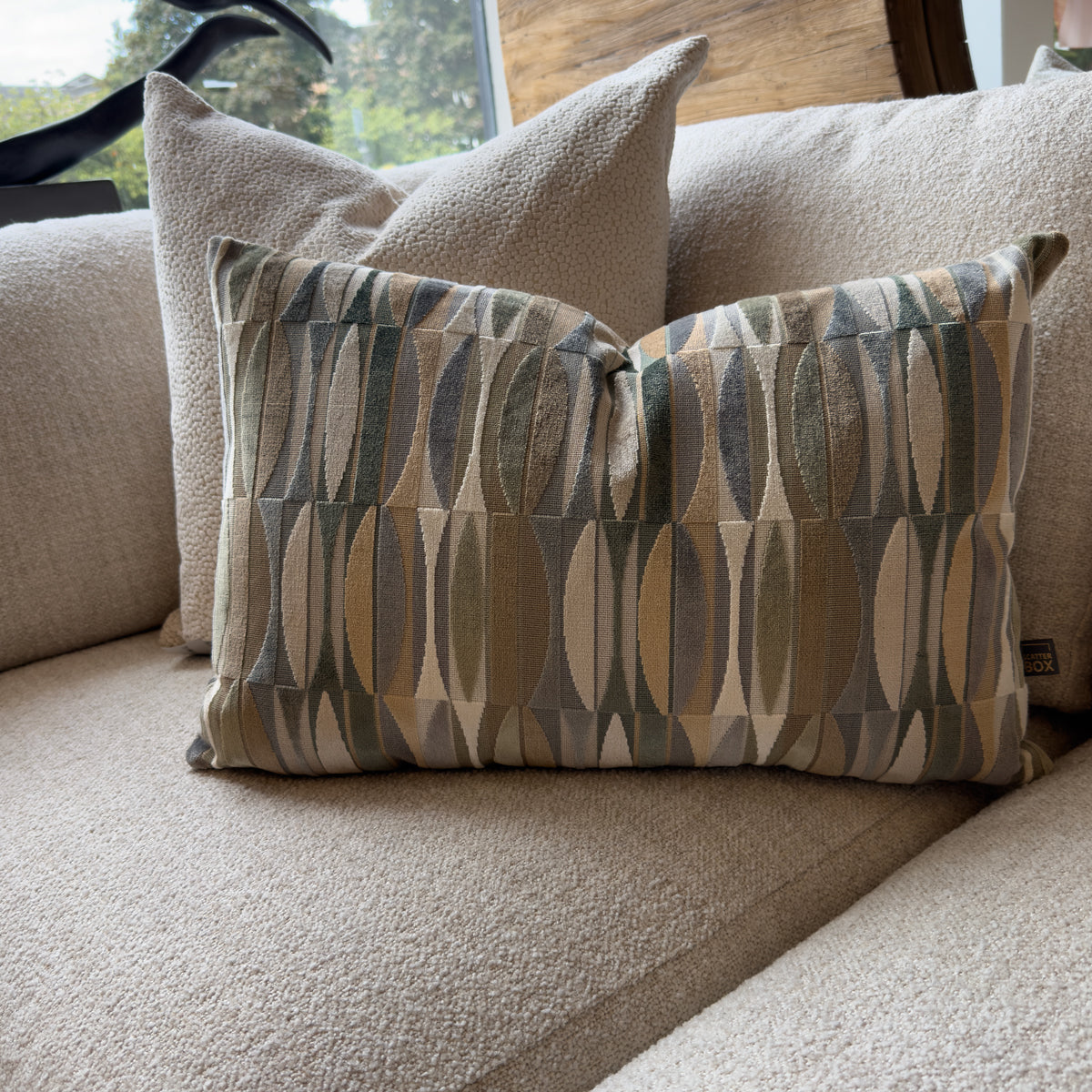 Close-up of a geometric throw pillow in muted beige, gray, and sage green on a cream sofa, creating a cozy, textured setting by a window.