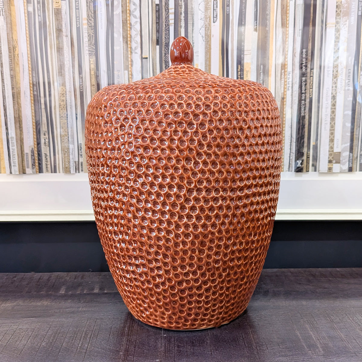 A deep orange lidded jar, sitting on a dark wood surface with an artwork of vinyl records in the background.