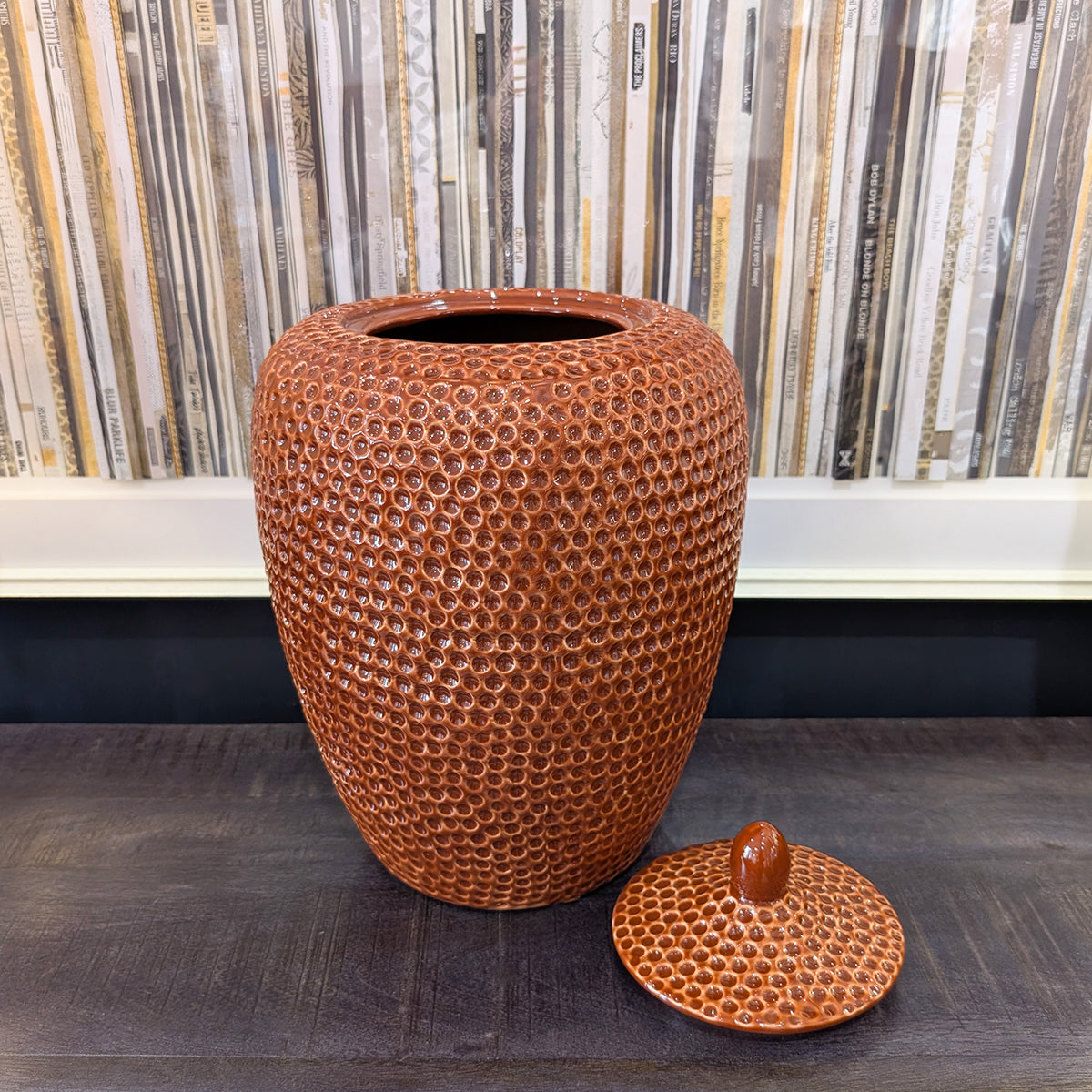 A rust dimpled jar with the lid off, sitting on a dark wood sideboard infront of a vinyl collection art picture.