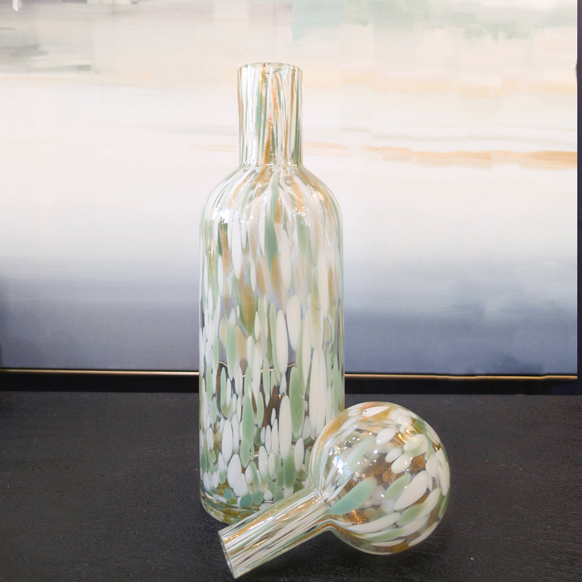 A sage green speckled lidded vase with its stopper removed, sitting on a black table top.