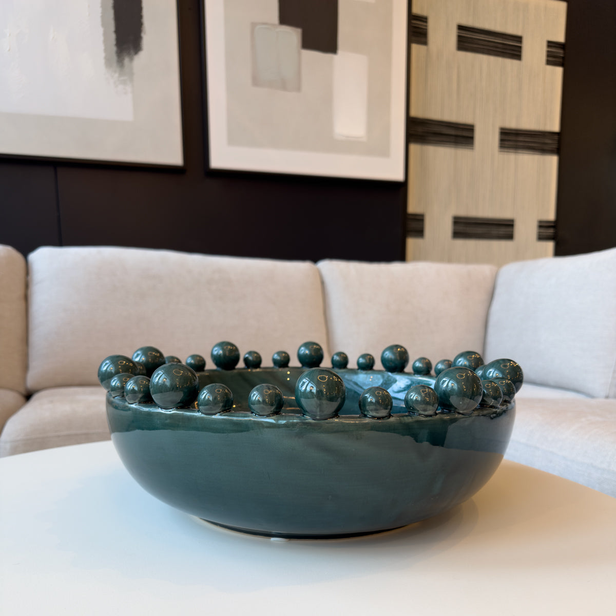 A teal bowl with bobble decorations, sitting in a cream living room with monochrome artwork on the walls.