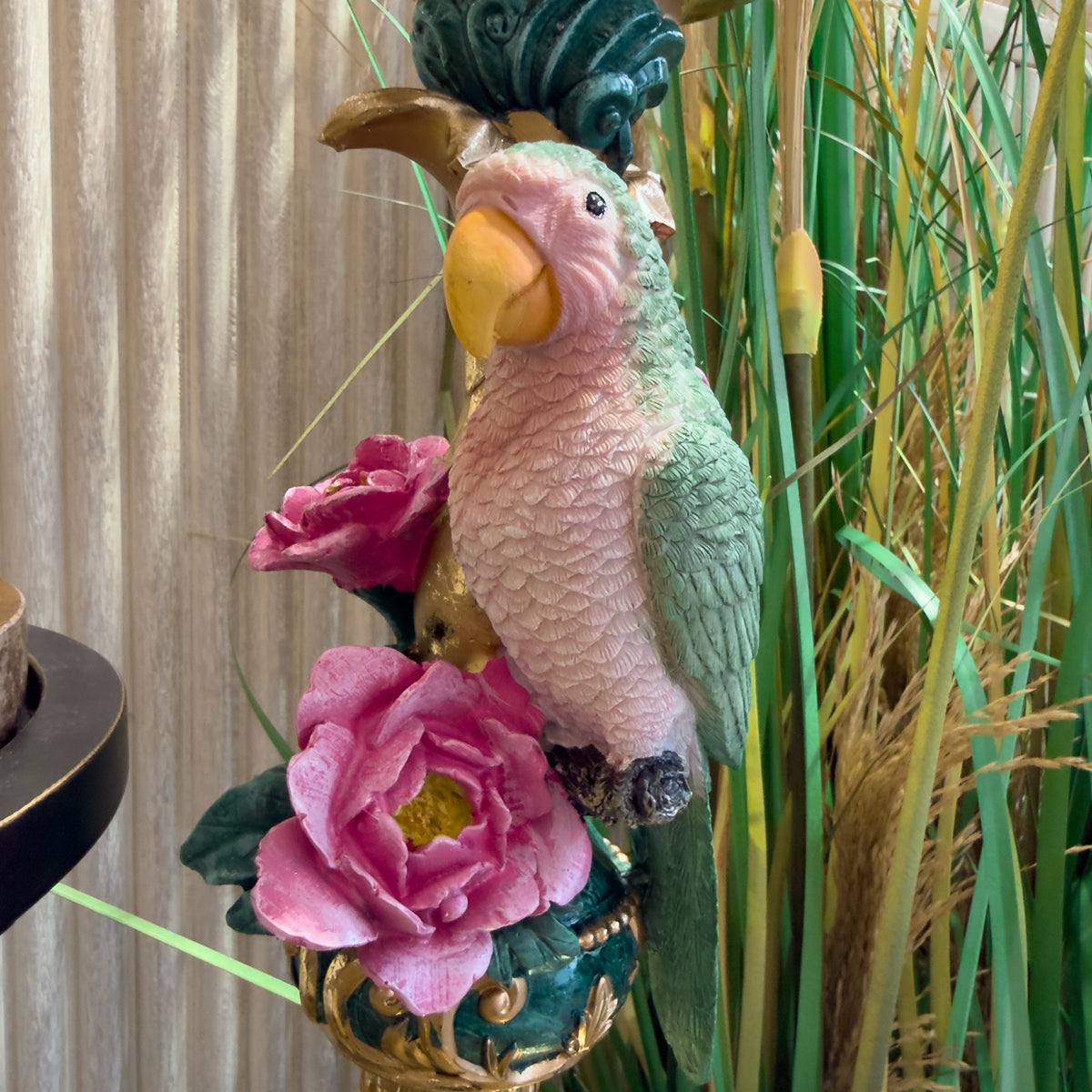 Ornate pink-and-green parrot candlestick with a curved beak, perched among vivid pink flowers and tall grasses.