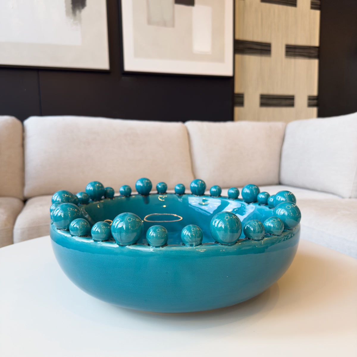 A shiny turquoise bowl with bobbles around the rim, sitting on a cream coffee table in a living room.