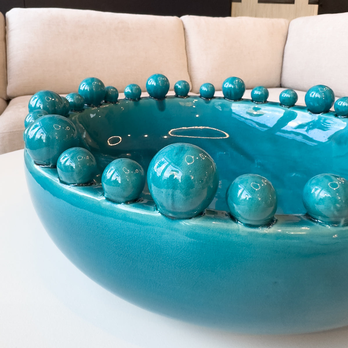 A close up of a high gloss blue bowl, sitting on a white coffee table infront of a cream sofa.