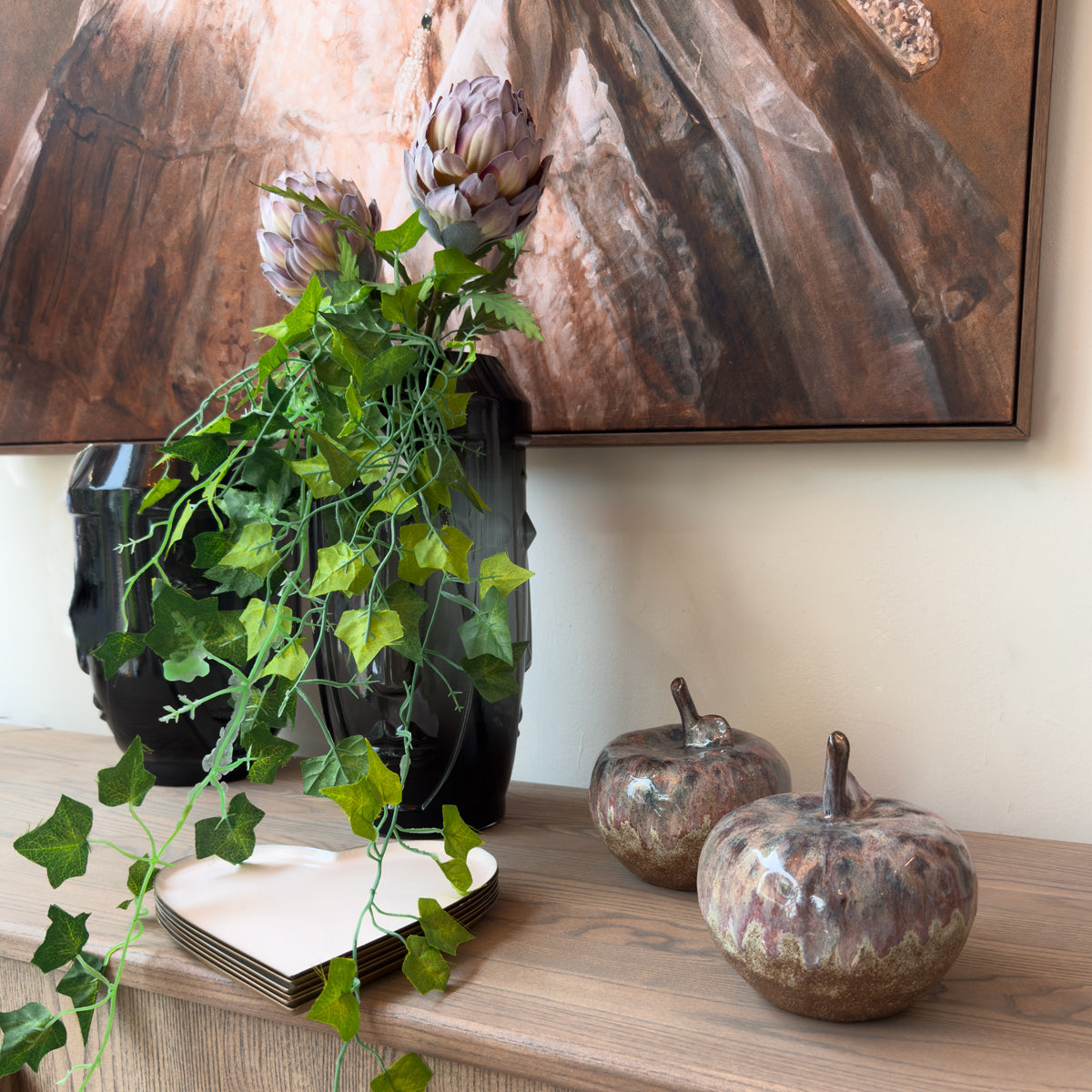Ivy and artichoke flowers, black vase, ceramic brown watercoloured mini ceramic apples, against a cream painted wall with an abstract painting on the wall.