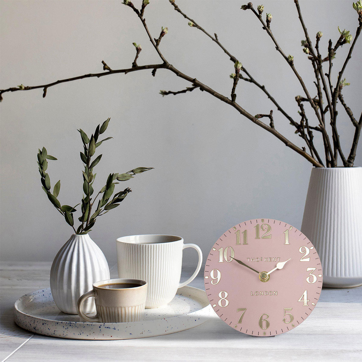Pink clock on a table with a speckled tray, mugs, and vase
