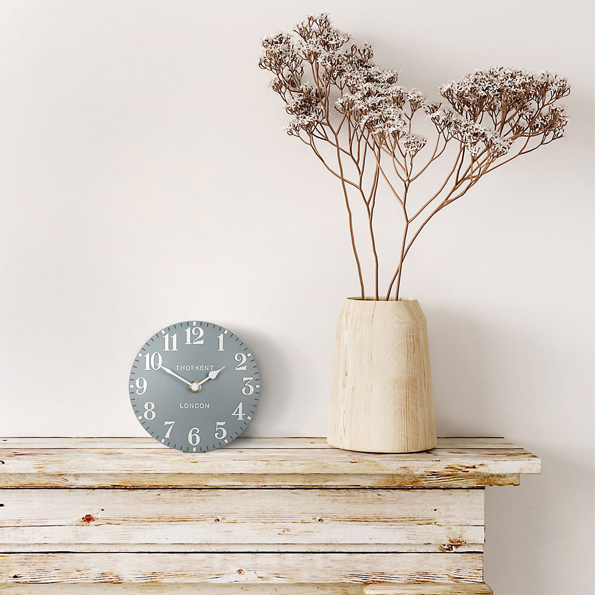 Clock and vase with dried flowers on a wooden surface against a white wall