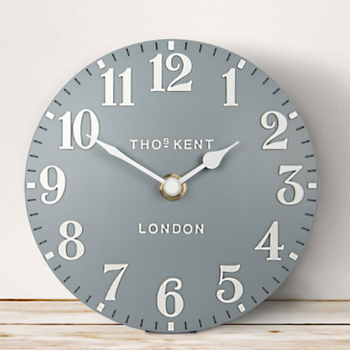 Grey blue wall clock with white numbers and 'Thomas Kent London' branding on a light wooden surface.