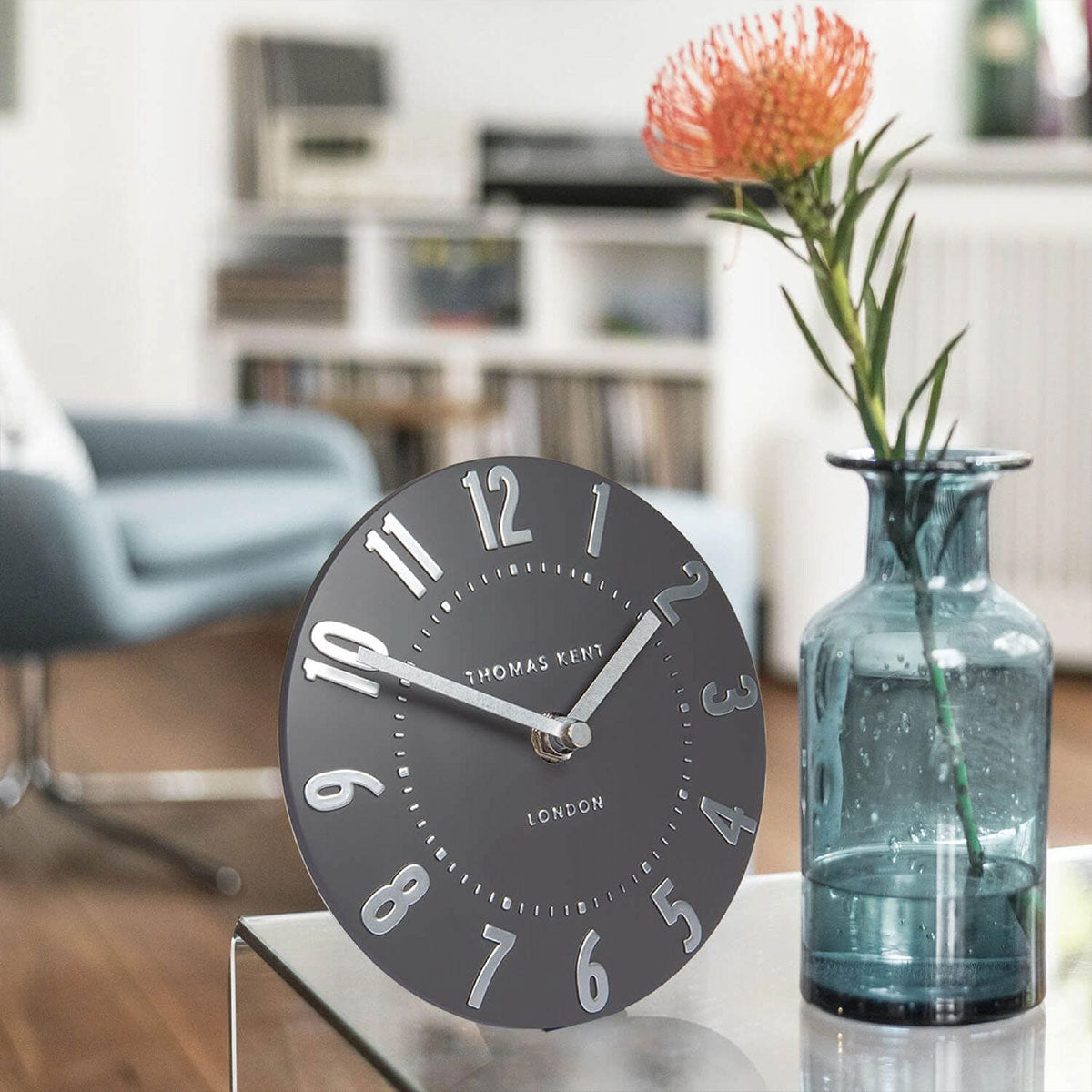 Round dark grey clock on a table with a vase of flowers in the background