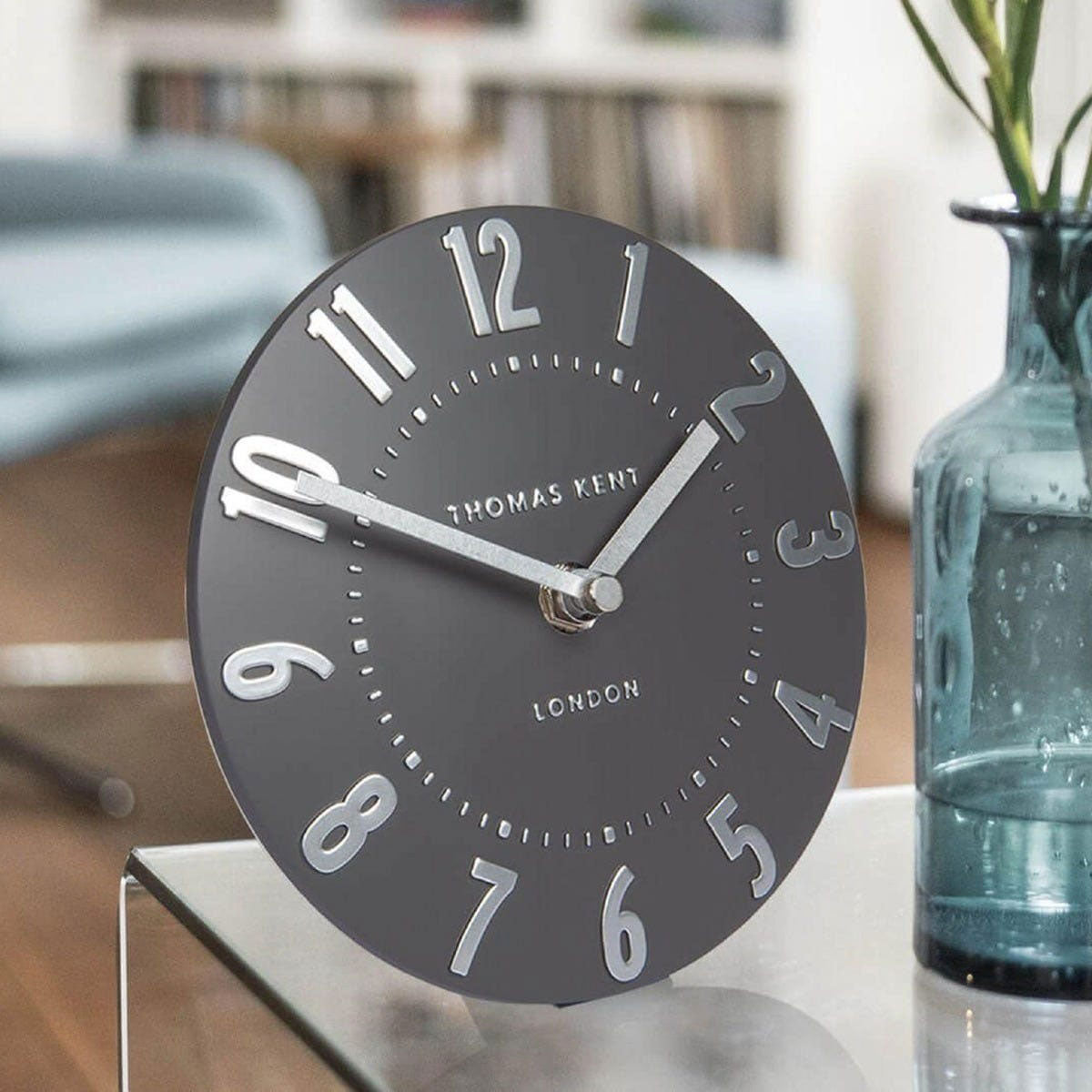 Dark gray clock with silver numbers and Thomas Kent branding on a stand, blurred background