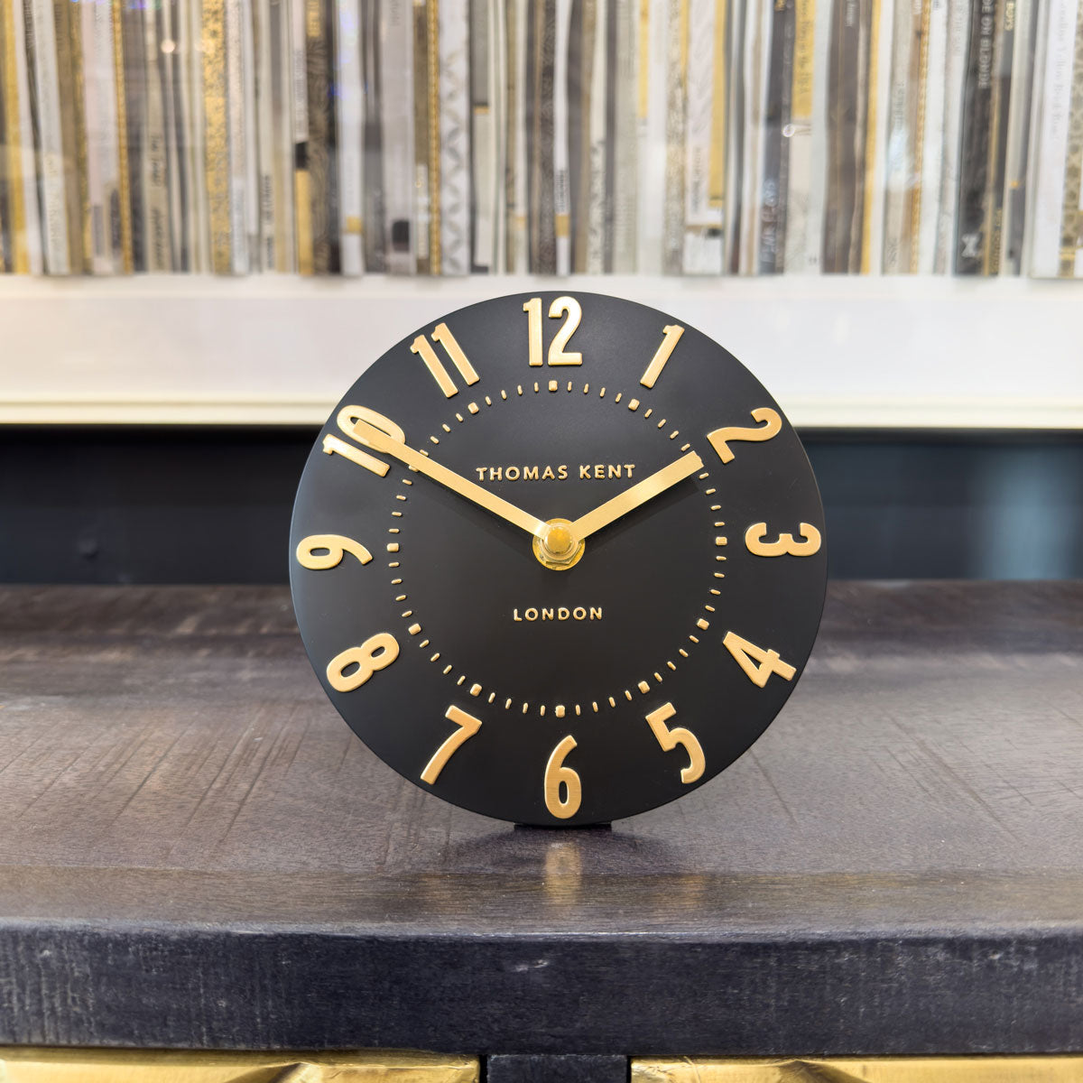 Black small clock with gold numbers and hands on a wooden surface, Thomas Kent brand visible