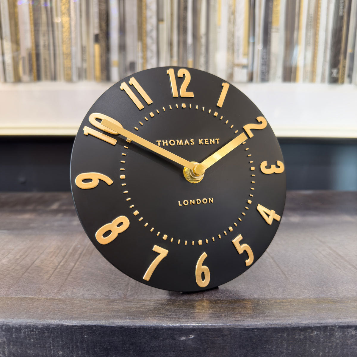 Black clock with gold numbers and hands on a wooden surface, branded 'Thomas Kent London'.