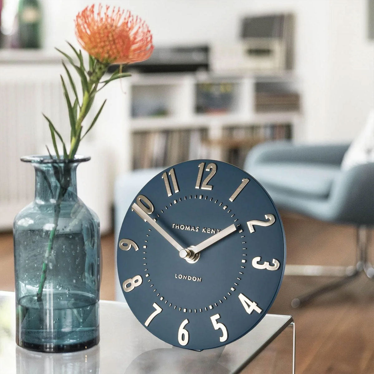 Blue clock on a table with a vase and flower in a blurred living room background