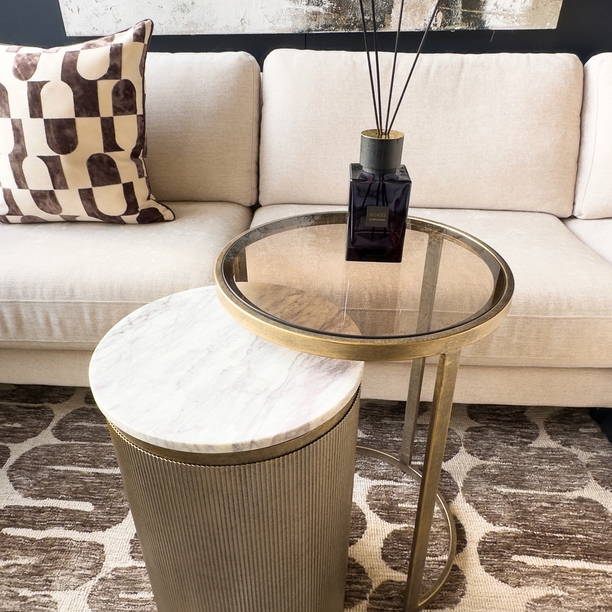 Round glass side table with gold frame next to a beige sofa with decorative pillows.