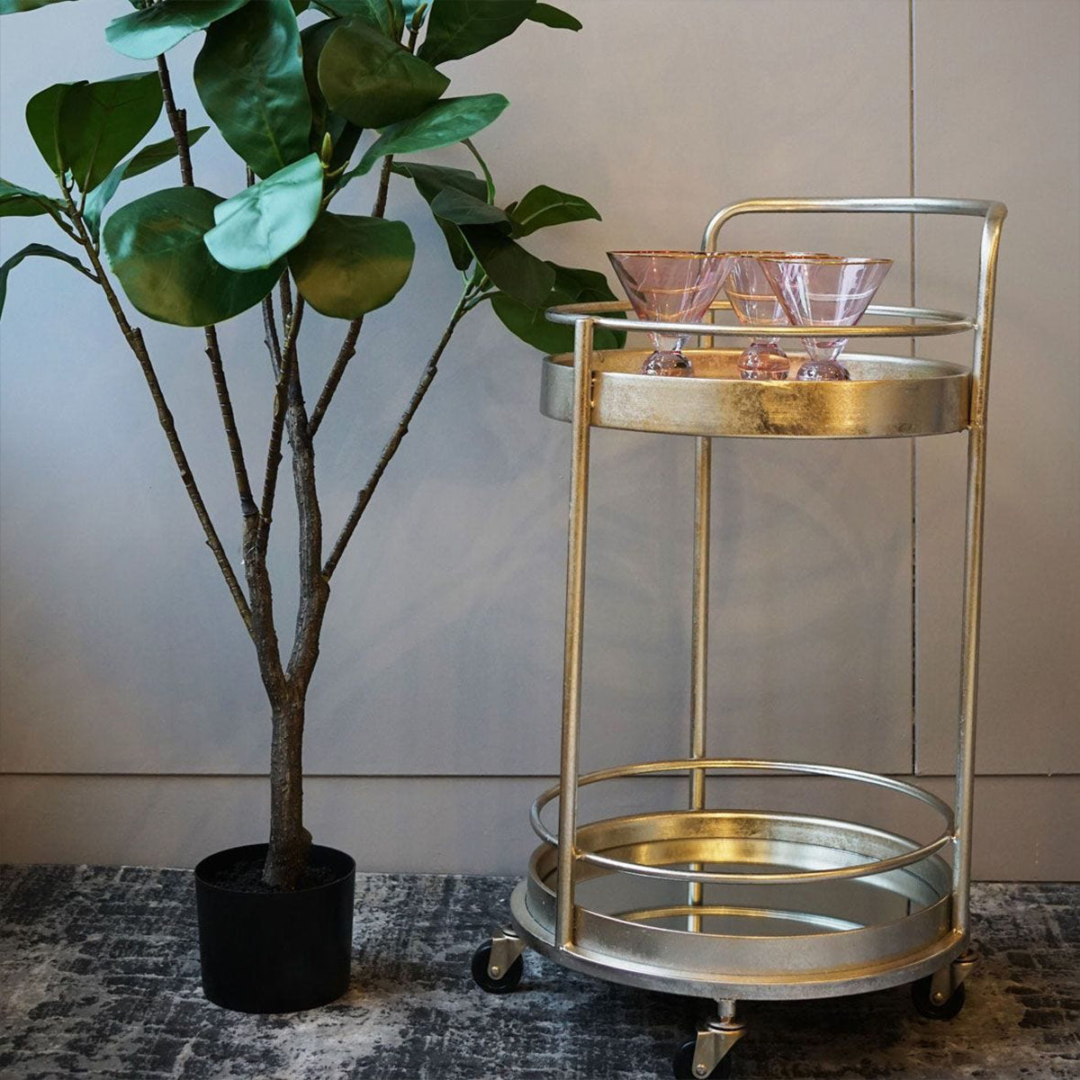 Antique silver small round bar trolley with mirrored glass shelves, positioned next to a potted plant with glasses on top and wheels on the base.