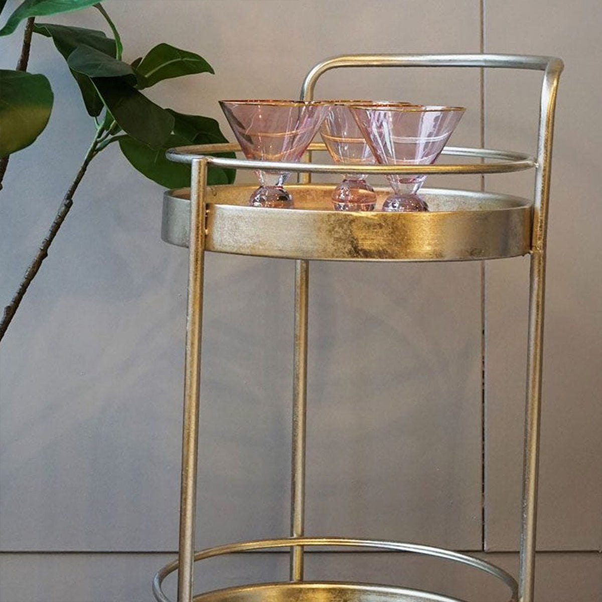 Gold bar cart with pink glassware against a neutral wall.