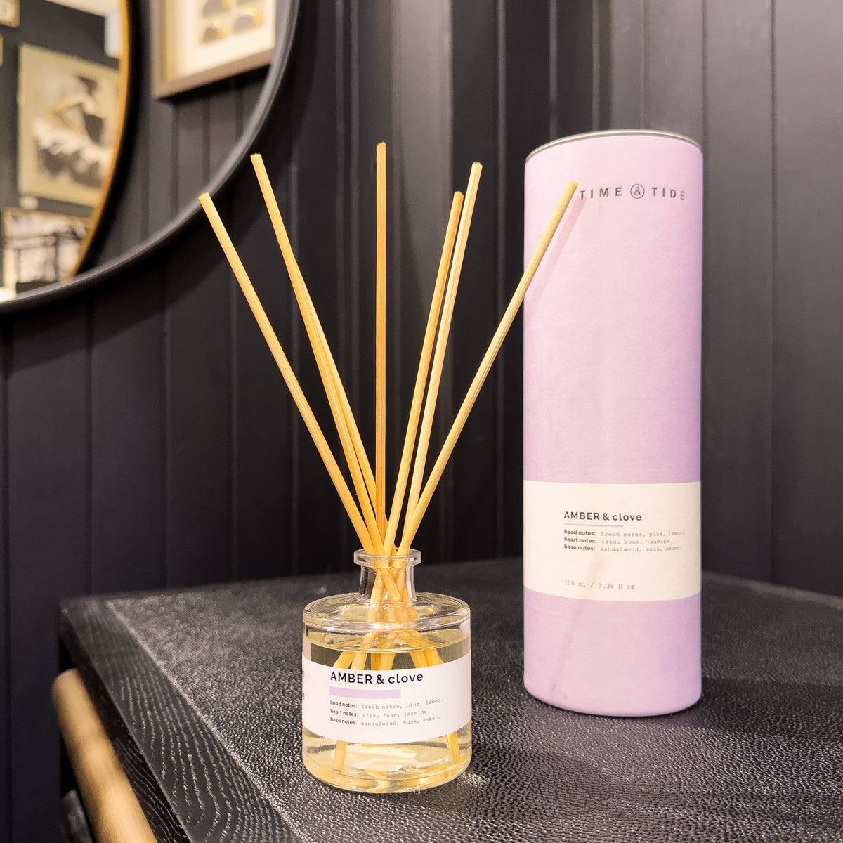 Diffuser with wooden sticks and a purple cylindrical container labeled 'Time & Tide' on a dark surface.