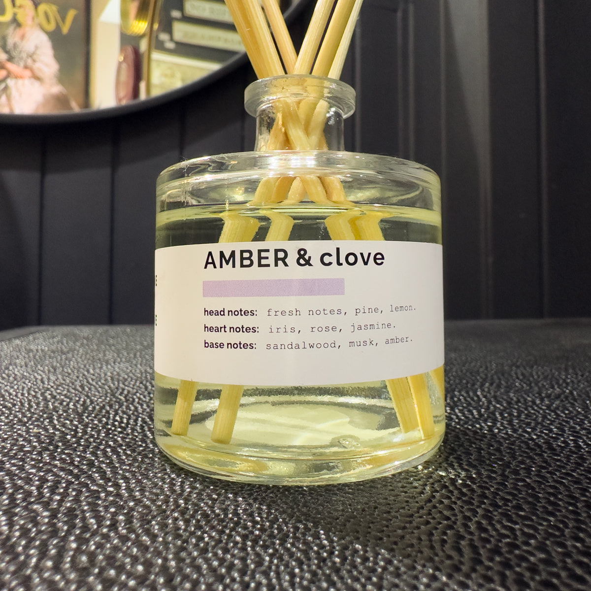 Reed diffuser labeled 'Amber & Clove' with wooden sticks in a clear bottle on a dark surface.