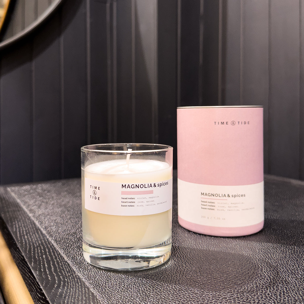 Candle and packaging labeled 'Time & Tide' with 'Magnolia & Spices' on a dark surface.