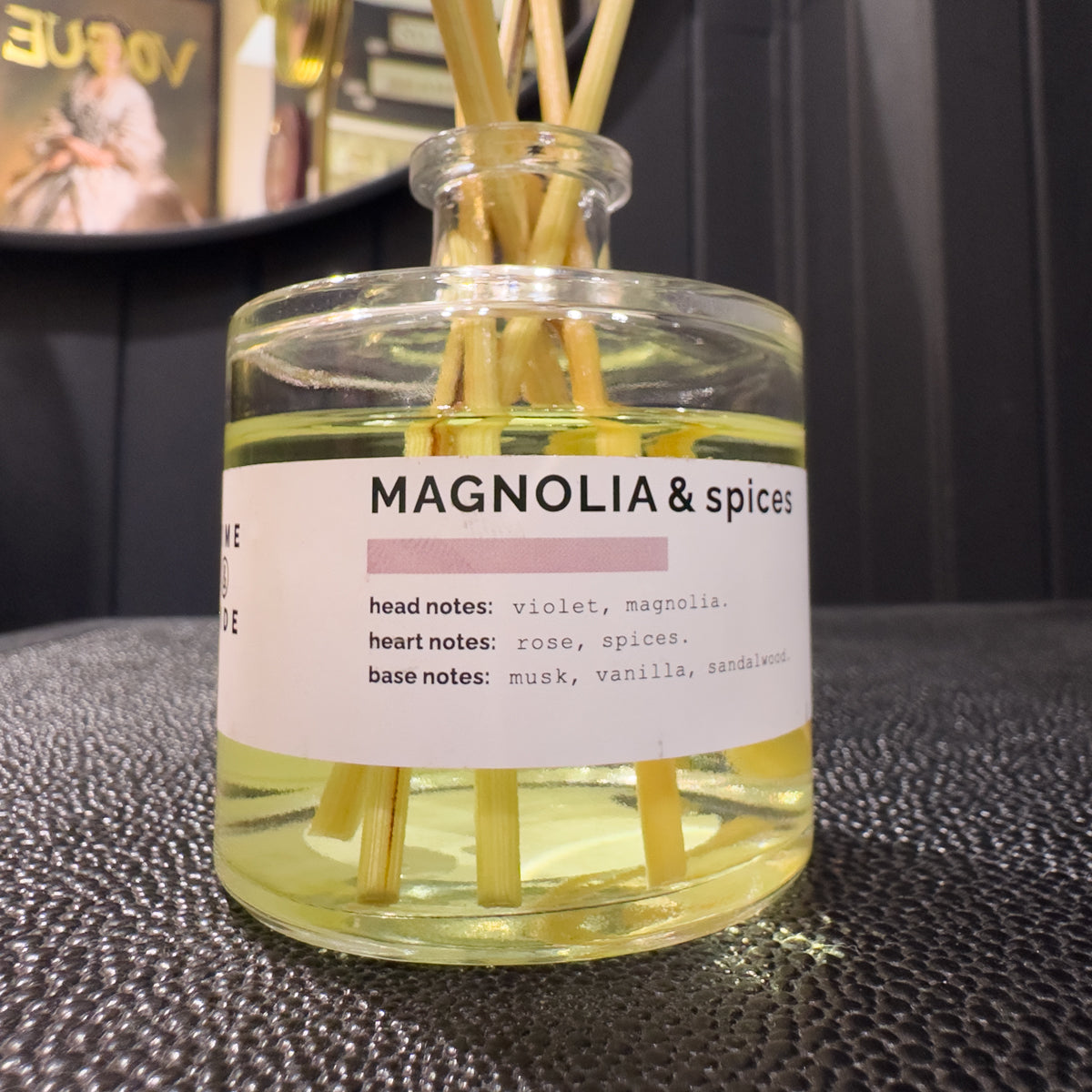 Glass diffuser bottle with sticks and a label reading 'Magnolia & Spices' on a textured surface.