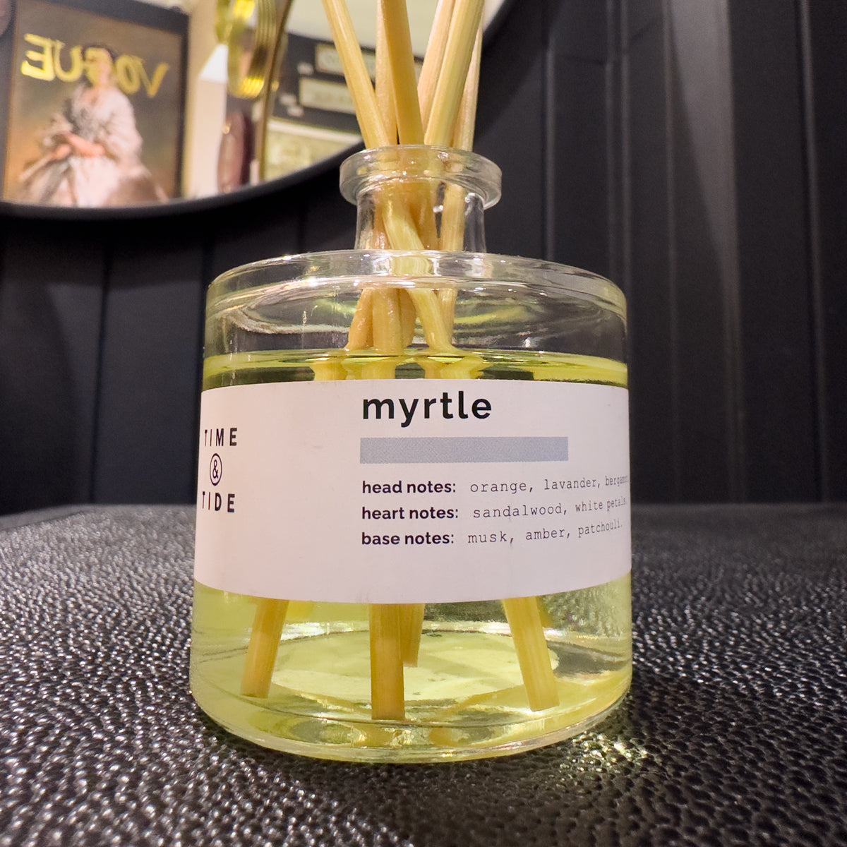 A glass diffuser bottle labeled 'myrtle' with wooden sticks inside, on a textured surface.