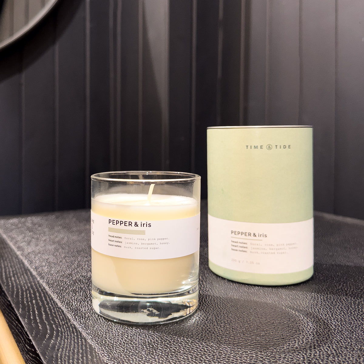 Two candles, one in a glass container and one in a green tin, on a dark surface with a dark background.