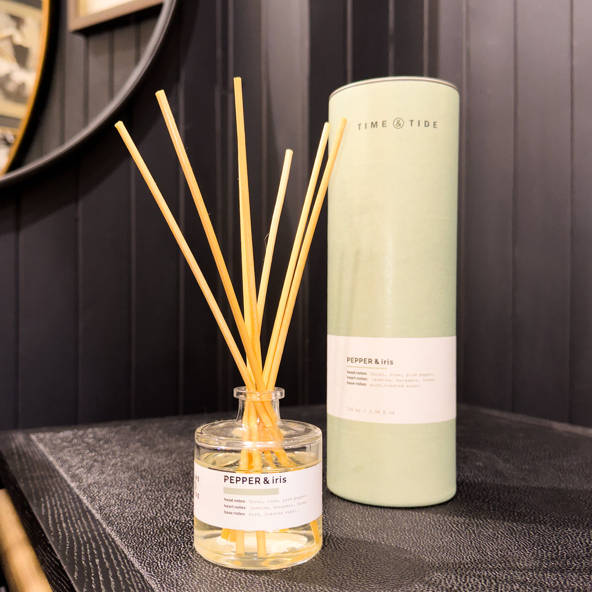 Reed diffuser with sticks and cylindrical container labeled 'Pepper & Iris' on a dark surface.