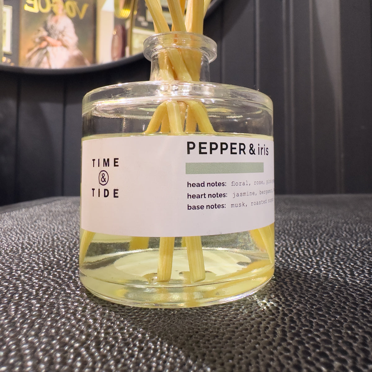 Clear glass reed diffuser with decorative sticks and 'Pepper & Iris' label on a textured surface.