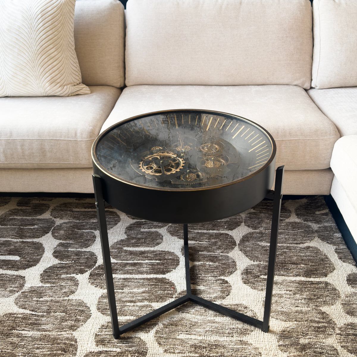 Round black gears clock side table with decorative glass top in front of a beige sofa on a patterned rug.
