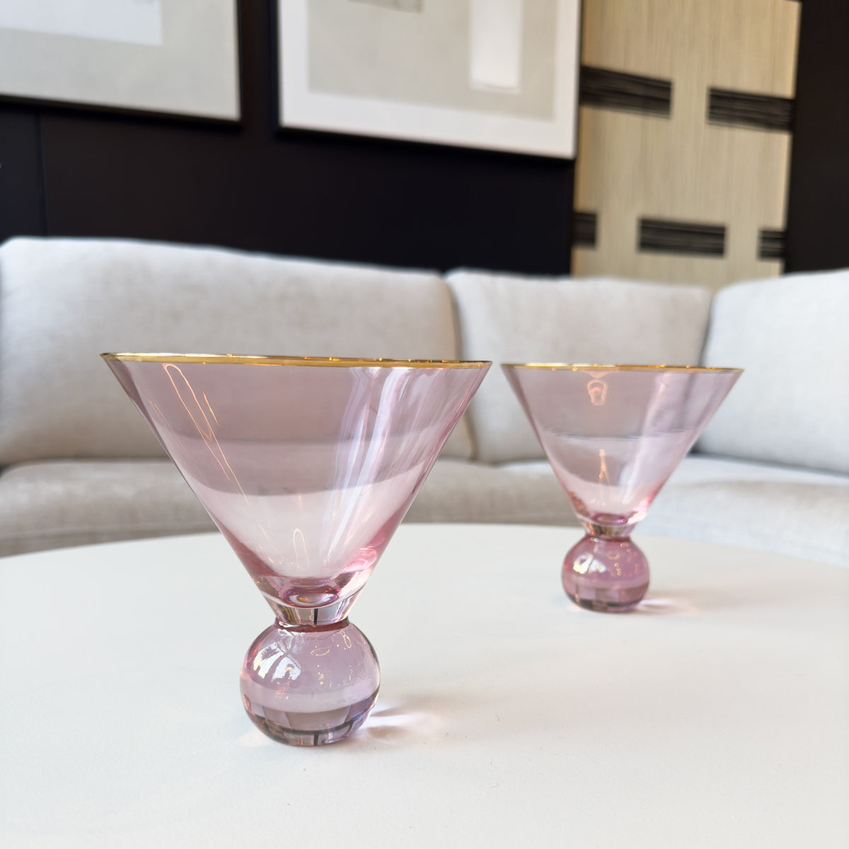 Two blush tinted martini glasses with gold rims and round glass feet on a table with a couch in the background.