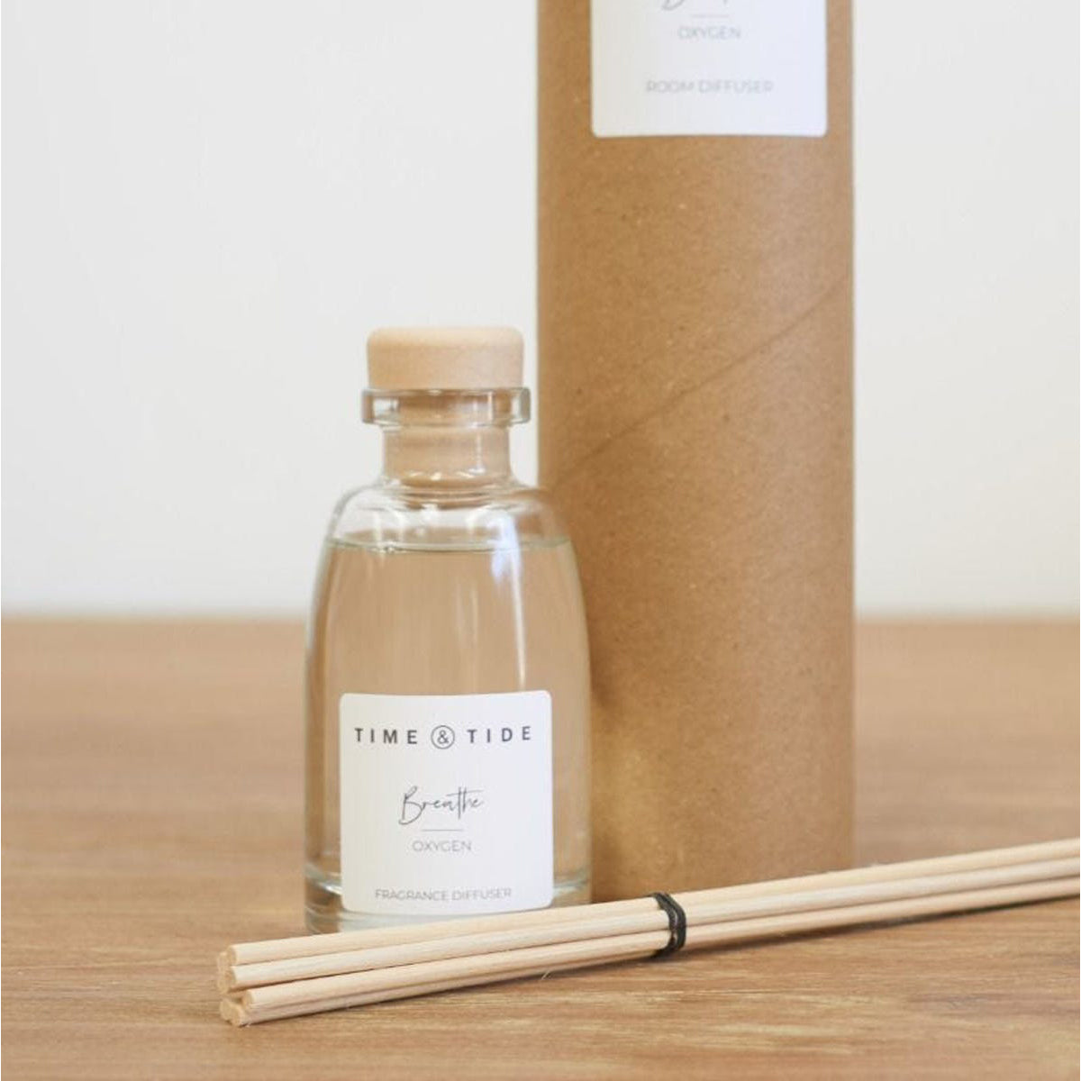 Clear glass diffuser bottle with wooden cap and reeds on a wooden surface, with a cardboard box in the background.