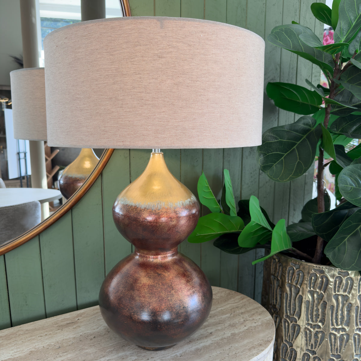 A warm copper and bronze table lamp with a wide beige shade sits on a pale stone console beside lush green plants, creating a calm, natural interior.