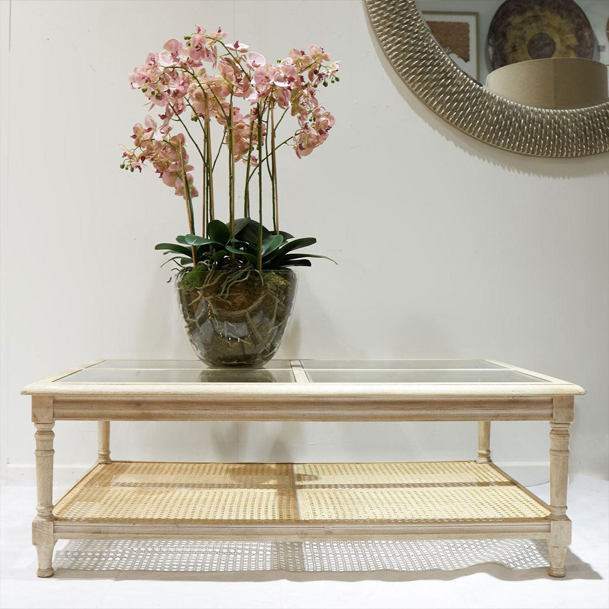 Rattan coffee table with a glass top and woven shelf, featuring a vase of pink flowers.