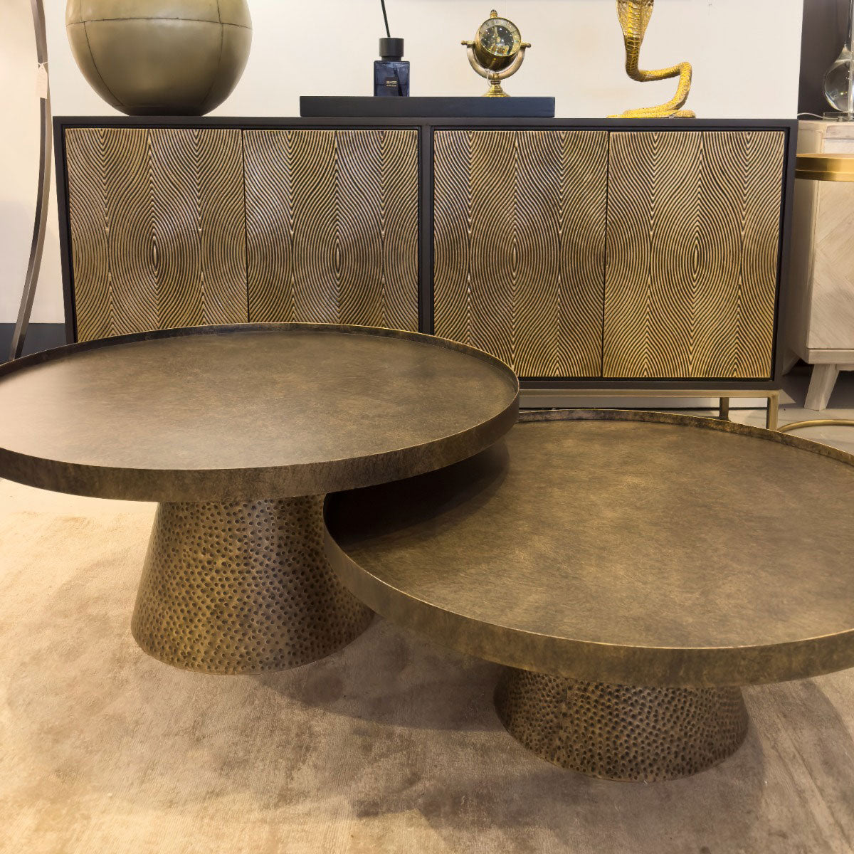Two round bronze tables with textured bases in front of a patterned cabinet.