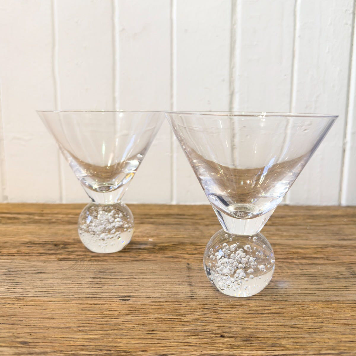 Two clear bubble base martini glasses on a wooden surface with a white background.