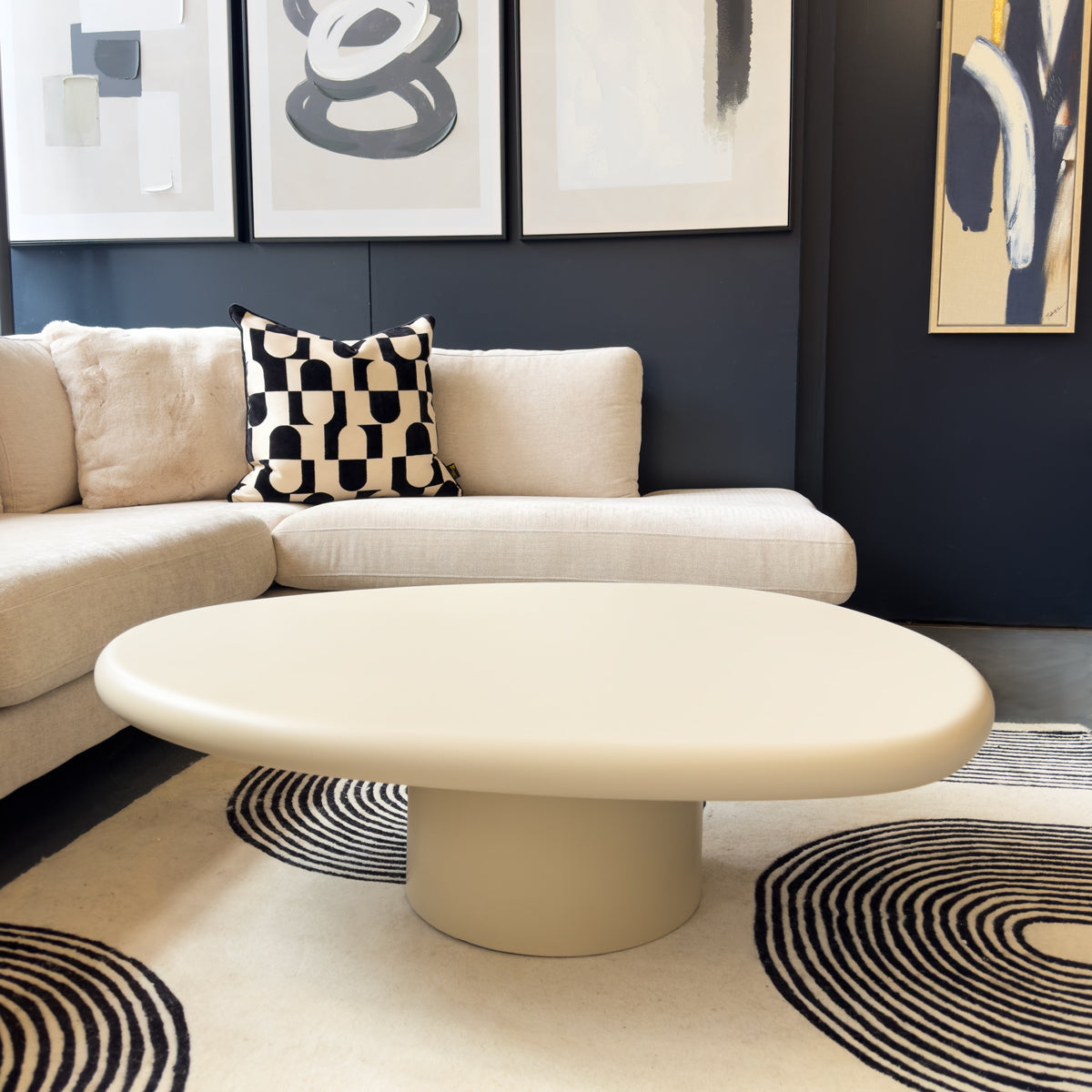 Beige sofa with a patterned pillow and a matching coffee table in a room with dark walls and abstract art.