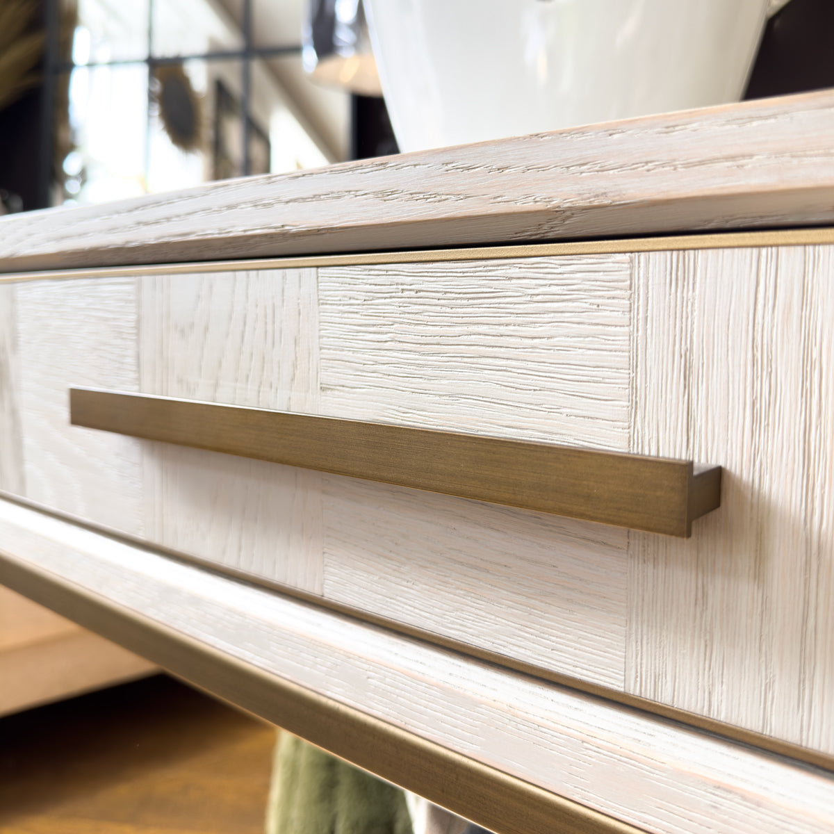 Close-up of a light wood dresser drawer with a rectangular brushed brass pull handle, showing the wood grain texture and a softly blurred interior background.