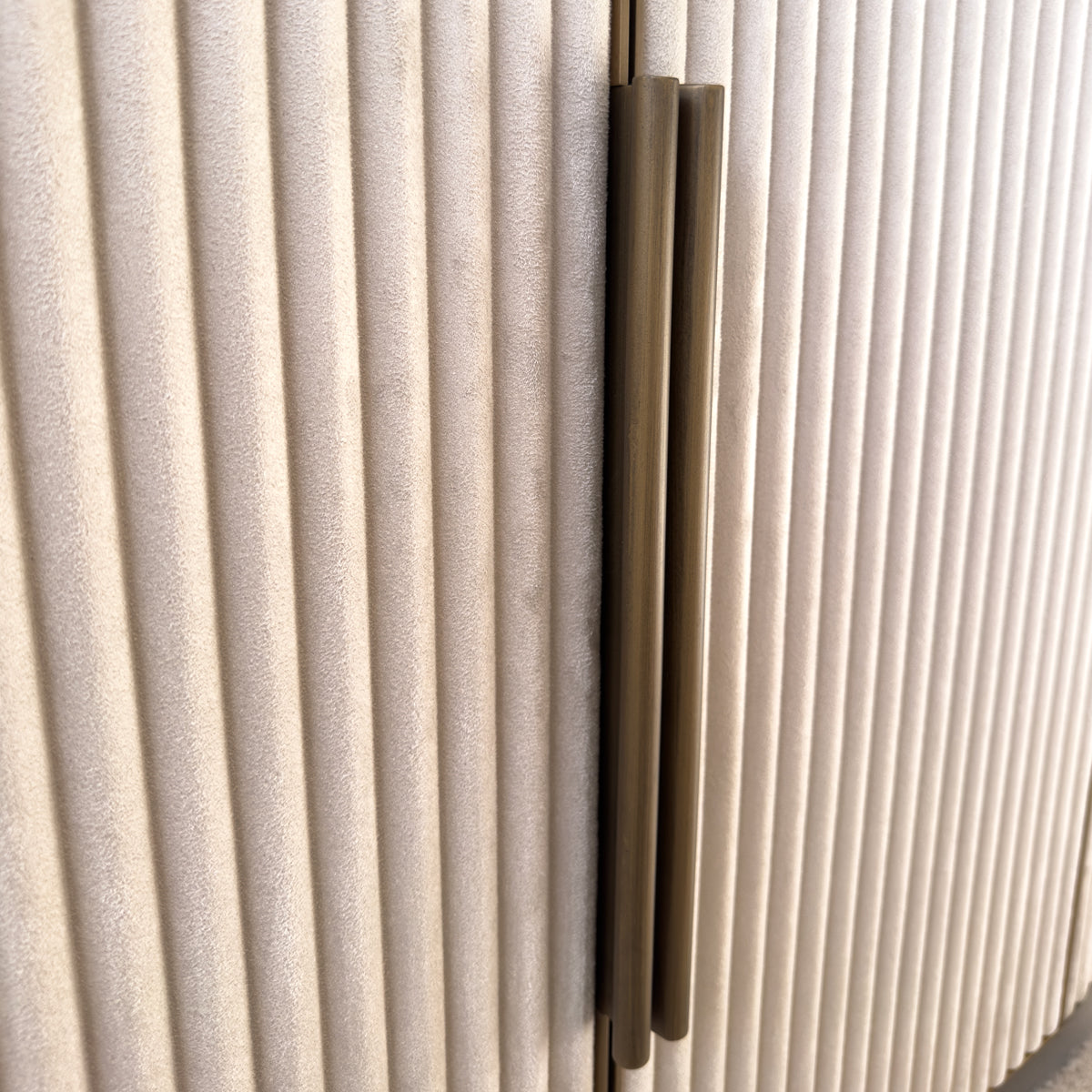 A close up of a neutral velvet sideboard with ridged doors and gold long handles.