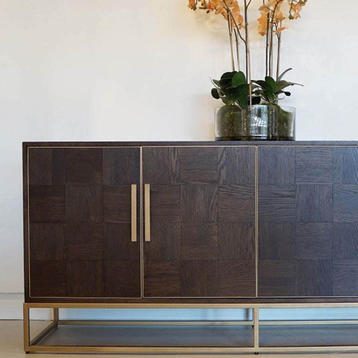 Dark wooden sideboard with gold handles and a vase of flowers on top against a white wall.