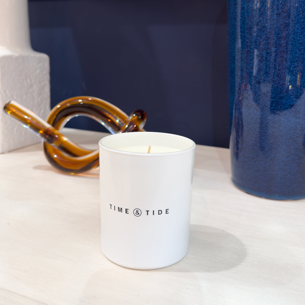 White jar candle with 'Time & Tide' branding on a white surface with a blue vase and brown object in the background.