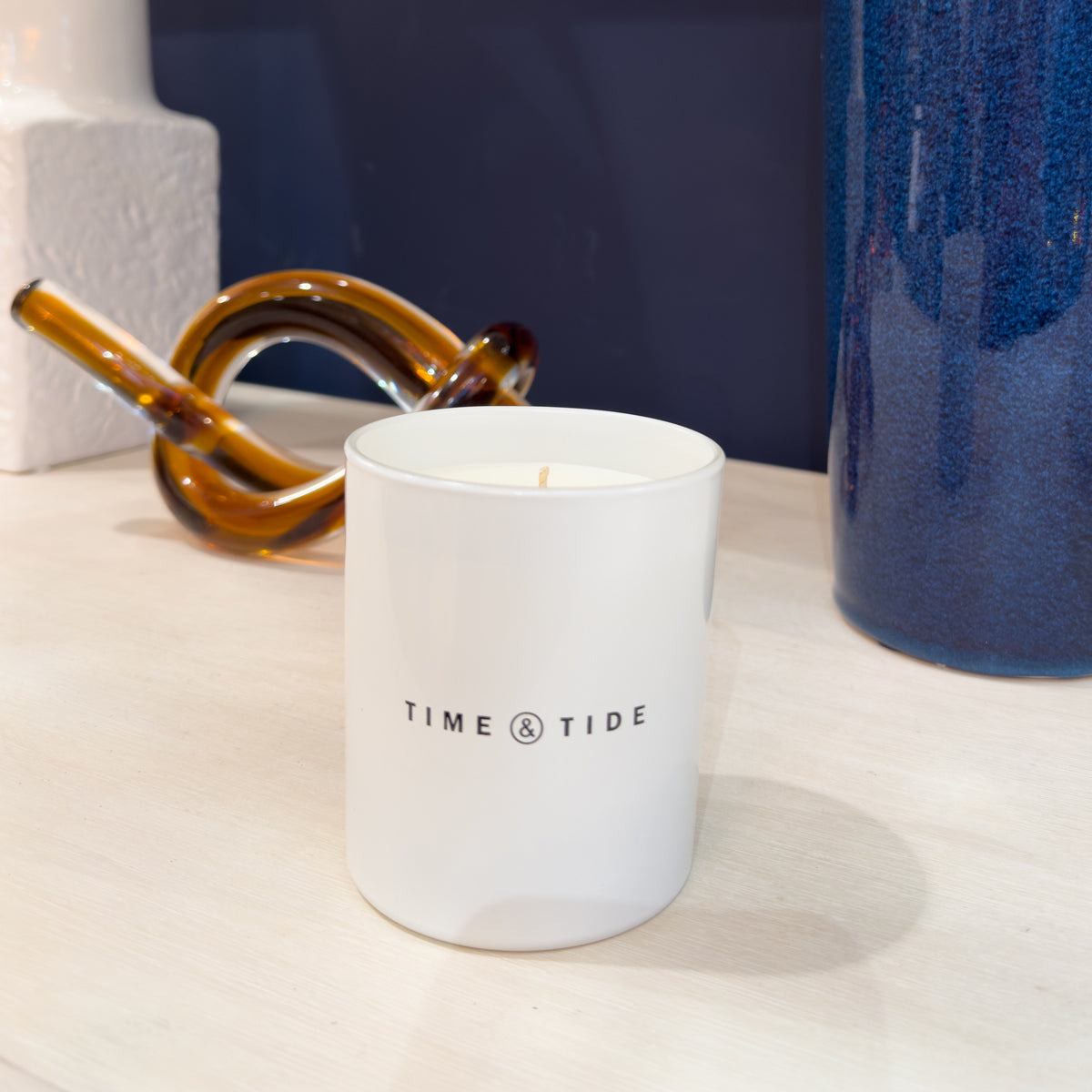White candle with 'Time & Tide' branding on a surface with decorative items.